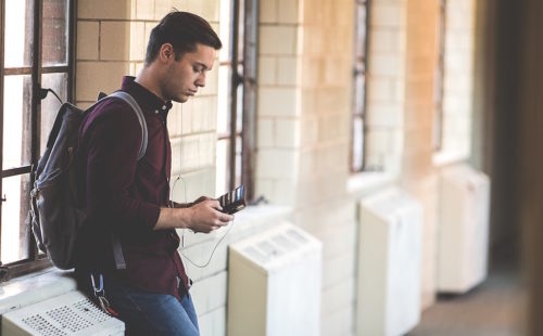 Teen with backpack uses phone while leaning against radiator.