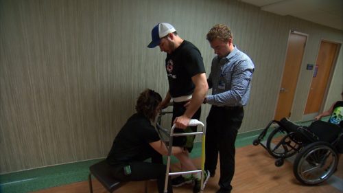 Paralyzed man walking with assistance.