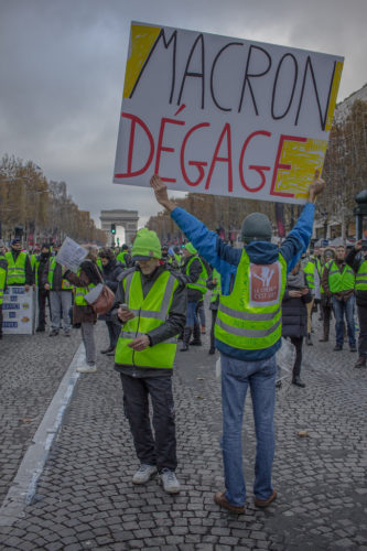 Protester holding a sign saying, Macron Degage, with Arc de Triomphe in background