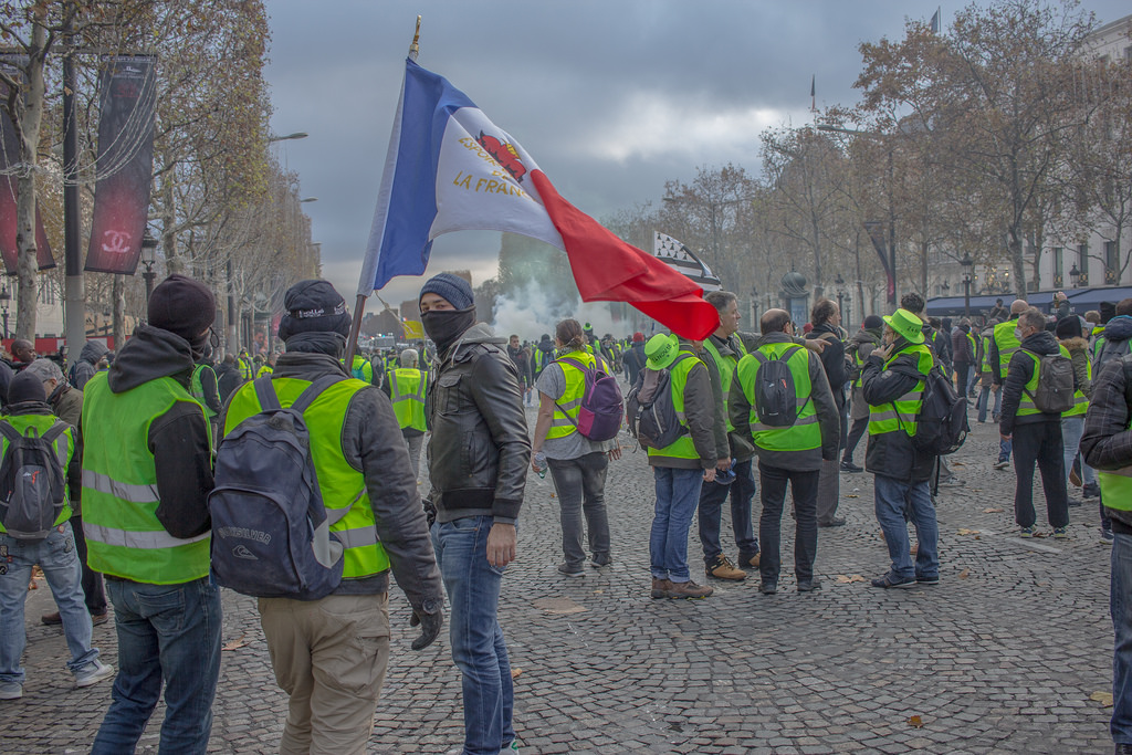 Masked protesters wearing yellow jackets and waving a flag are seen. Tear gas is seen rising in the distance.