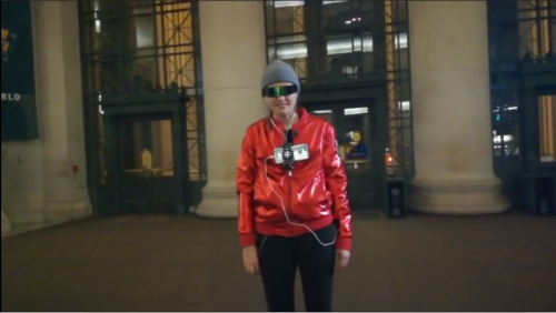 A woman in a red coat and dark glasses wearing a chest-mounted camera.