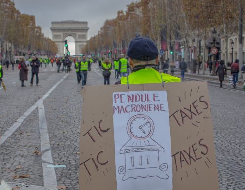 Man carrying a sign with a clock and the words "Tic-taxes, tic-taxes", with the Arc de Triomphe in the background.