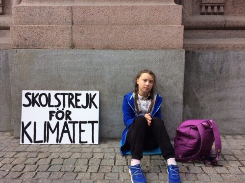 Greta seated with backpack. On the left is her sign in Swedish, which says, "School Strike for Climate".