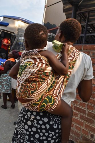 A little girl is in a wrapper on her mothers back. Just before she was picked up she had been crying. a stranger gives the girl a $2 bond note in the midst of a cash crisis to quite her and tells her she can buy her self something nice.