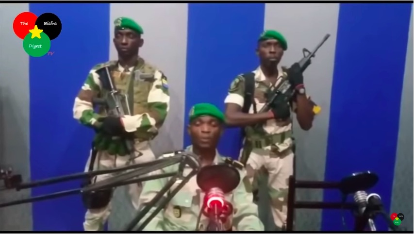 Three soldiers attempting a coup face the camera. One reads the message. Behind are two others with automatic weapons.