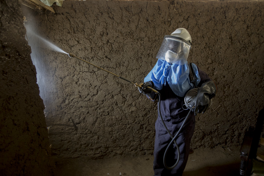 A woman in protective gear sprays a house for mosquitoes. The sprayer, Caroline Obinju Ocholla, is 33 yrs old, married with 4 children - 5,8,10, and 12 years old. She currently volunteers as a CHV. Photo by Jessica Scranton/AIRS