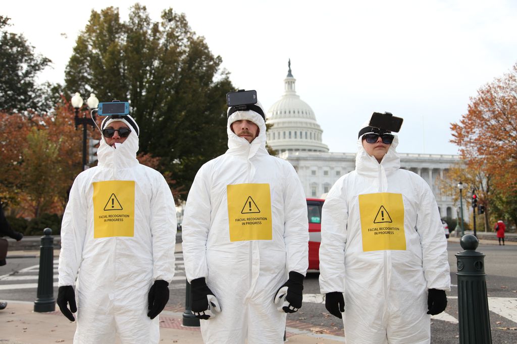 Three activists in white hazmat suits with cell phones strapped to their heads scan people in Washington, DC.