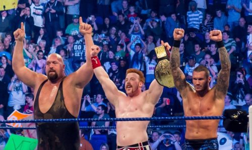 Three professional wrestlers raise their arms before a crowd.