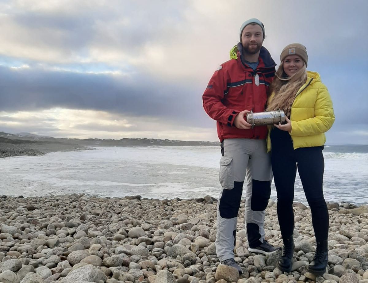 Conor McClory and Sophie Curran standing on a beach holding the time capsule.