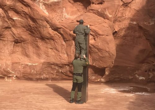 A worker stands on another worker's shoulders to look at the top of the metal monolith found in the Utah desert.