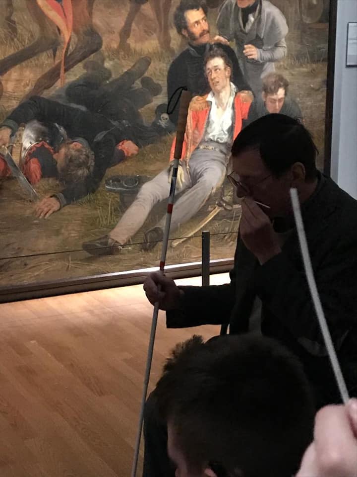 A man with a white cane seated in front of a painting sniffs a scented cardboard strip. In the foreground, another strip is visible, close up. Hannes walrafen smell tour for the blind at Rijksmuseum.