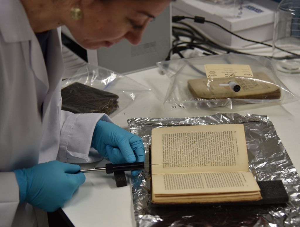 The smell of an old book is being extracted in the Heritage Science Lab at UCL. A gloved-handed scientist pulls a plunger from a tube pointed at an old book that is lying open.
