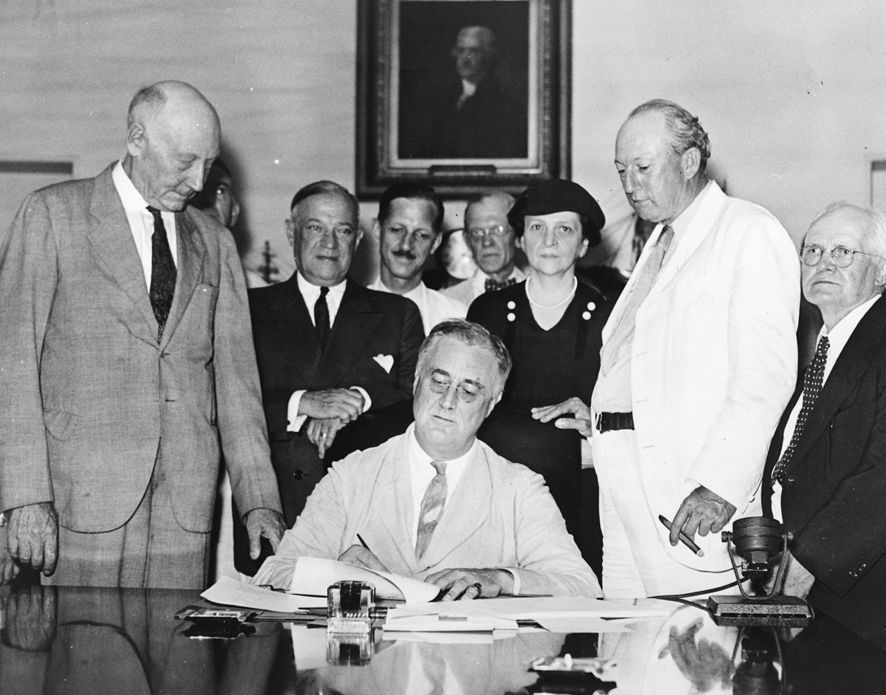 President Roosevelt signs Social Security Act as several politicians, including Frances Perkins, look on.