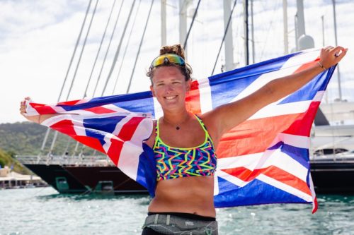 Jasmine Harrison stands in front of a sailboat holding the UK flag stretched out behind her.