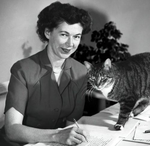 Black and white image of Beverly Cleary, around 1955. She's smiling at the camera while appearing to edit a typewritten manuscript with a pencil. A cat is beginning to walk across the manuscript.