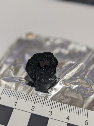 A carbonaceous chondrite meteorite found in the UK. A measuring tape shows it as being about 2.5 centimeters in diameter.