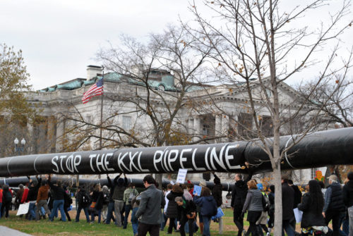 People protesting against the Keystone XL pipeline hold up a large pipe that says "Stop the KXL Pipeline" in front of the White House.