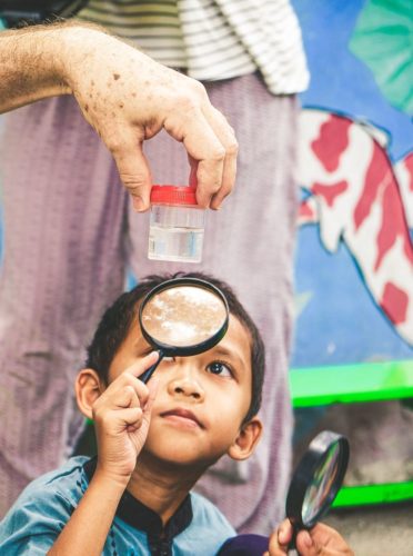 A boy examines a liquid-filled container with a magnifying glass. Community release of Wolbachia treated mosquitoes in Yogyakarta, Indonesia.