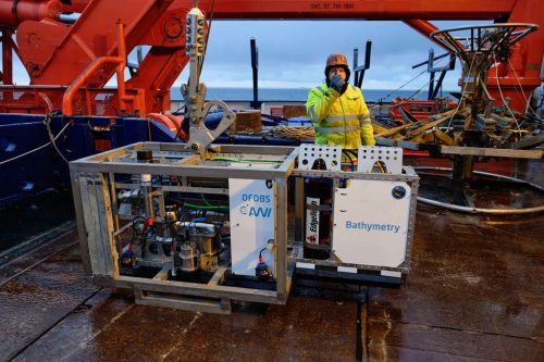 The Ocean Floor Observation and Bathymetry System (OFOBS) before deployment. A man in a hard hat and reflective yellow jacket stands behind the large, crate-sized OFOBS on the wet deck of a ship.