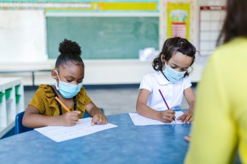 Two young girls sit at a table writing, wearing masks in school.