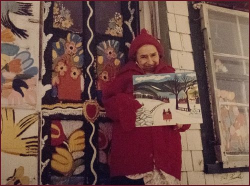Maud Lewis holding a painting in front of a house.