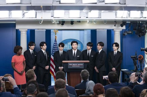 BTS members speak with reporters at the White House. The seven members, wearing black suits, stand behind the White House podium next to the White House press secretary.