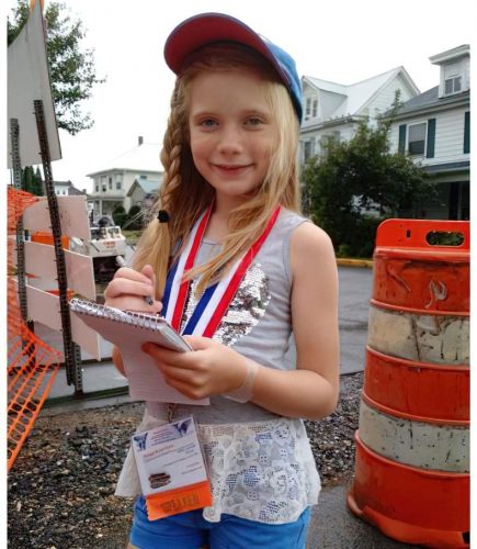 Hilde Lysiak wearing a baseball cap and a press badge glances at the camera while writing in a reporter's notebook.