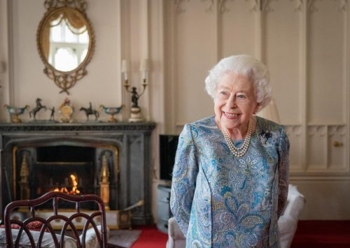 A photo of a smiling, white-haired Queen Elizabeth II wearing a blue paisley dress and pearls, standing in front of a fire.