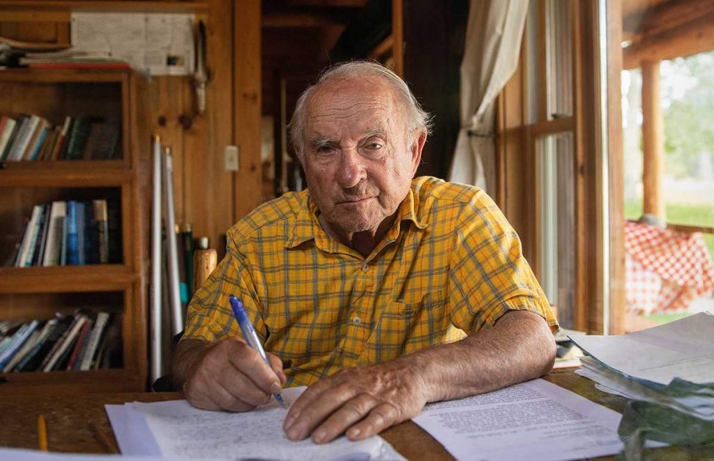 Yvon Chouinard in a simple wood-paneled room looks at the camera with pen in hand as he is writing.