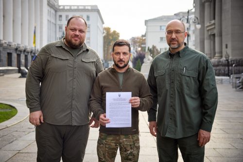 Ukraine President Volodymyr Zelenskyy standing between two other men holds a paper representing Ukraine's application to join NATO.