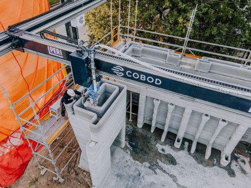 A high angle shot looking down on a worker and the massive 3D printer as it works on the second story of the house.