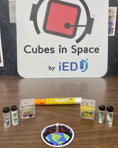 An EpiPen, with two cubes and two vials for each cube on display in front of a sign saying "Cubes in Space".