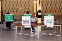 A man oversees two ballot boxes on a table during the 2023 general election in Thailand. In the background, voters are filling out ballots.