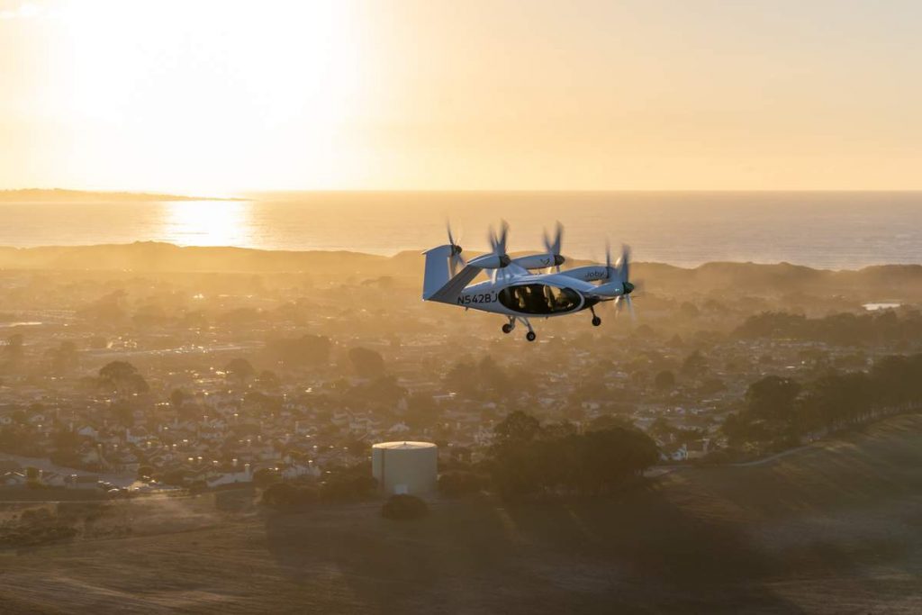 Joby Aviation's aircraft in the air above a city with the sea in the background.