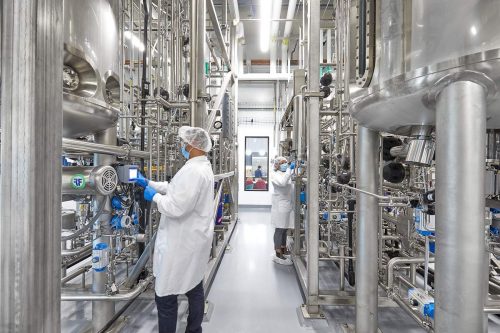 Cultivation room at Upside Foods. Two employees wearing hairnets, face masks, and lab coats are checking readouts on LCD panels in a complicated network of stainless steel pipes and tanks.