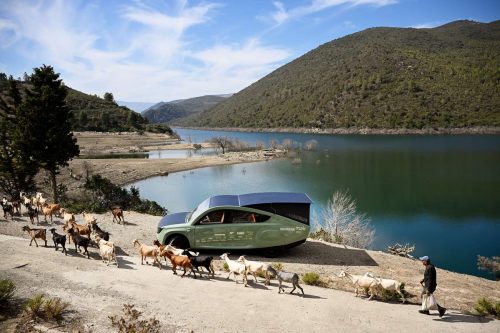 The Stella Terra solar car is seen charging by the side of a lake, with its solar panels raised, while a flock of goats walk past.