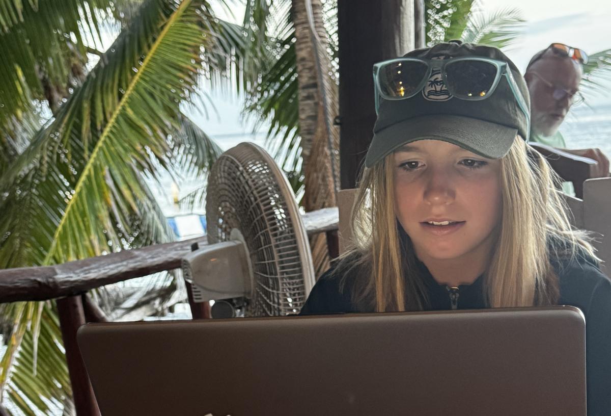 Mia DaPonte doing an online test for a diving course. She can be seen looking at a laptop screen, with the ocean and palm fronds in the background.