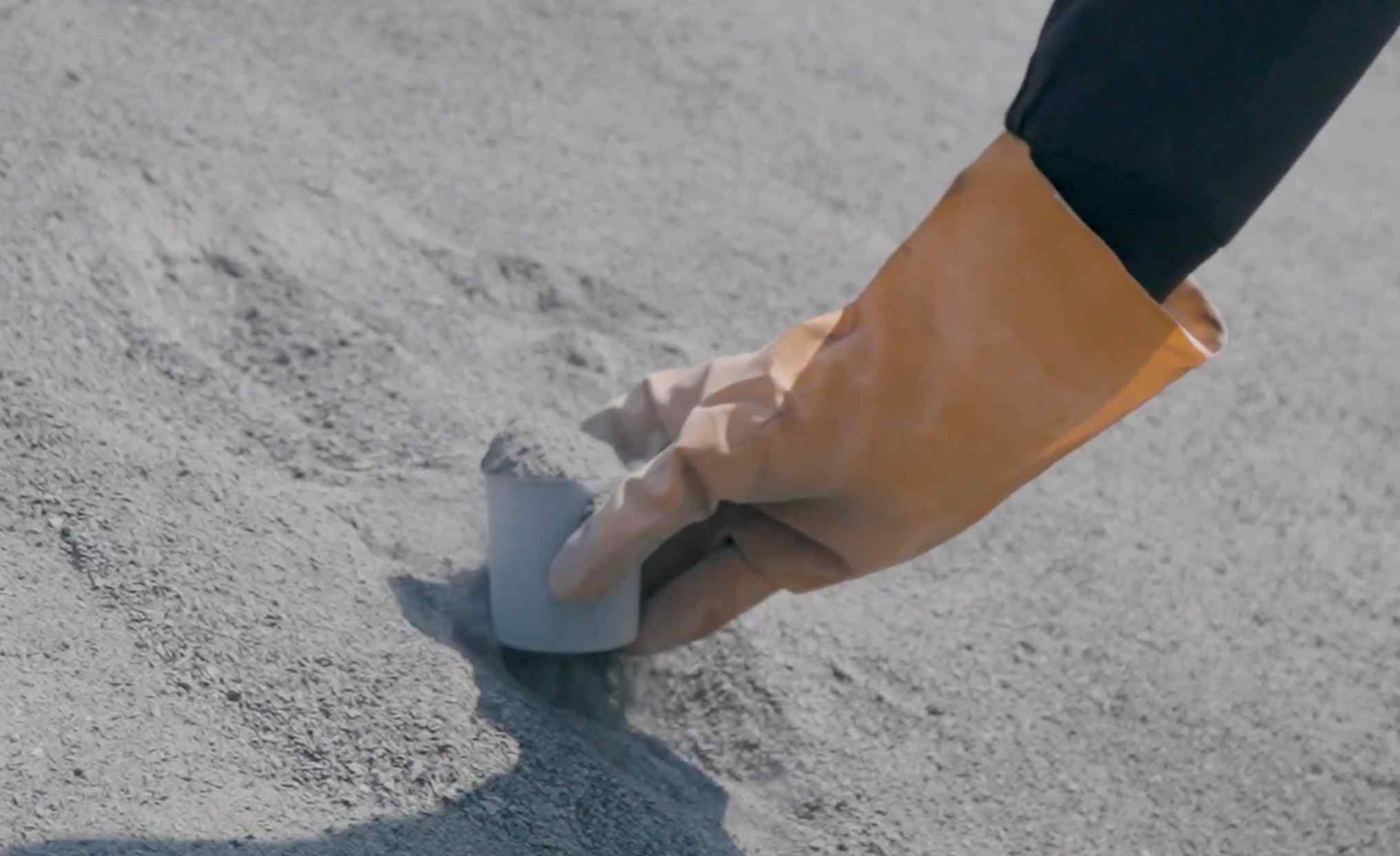A yellow rubber gloved hand scooping up a cup full of basalt dust from a huge pile.