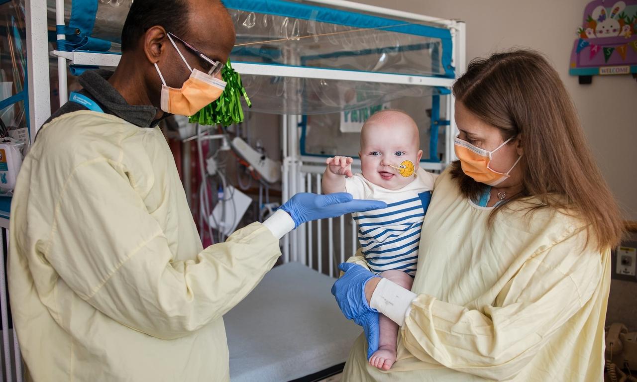Two masked doctors in medical gowns with the baby, KJ, in a striped shirt. A female doctor on the right holds KJ while the baby reaches out for the gloved hand of the Black male doctor on the left.