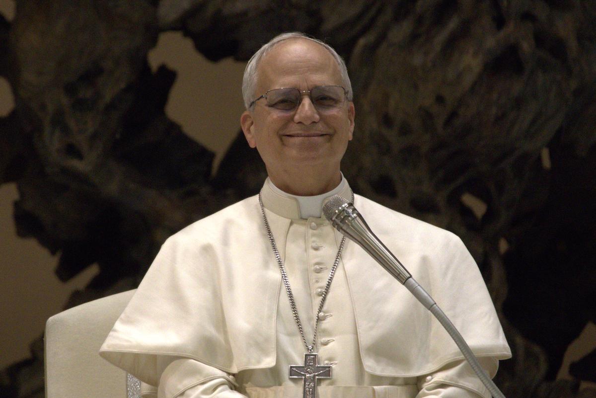Pope Leo XIV, dressed in white and wearing glasses smiles during an audience with the media on May 12, 2025. He wears a heavy silver cross on a chain around his neck. A microphone is in front of him.