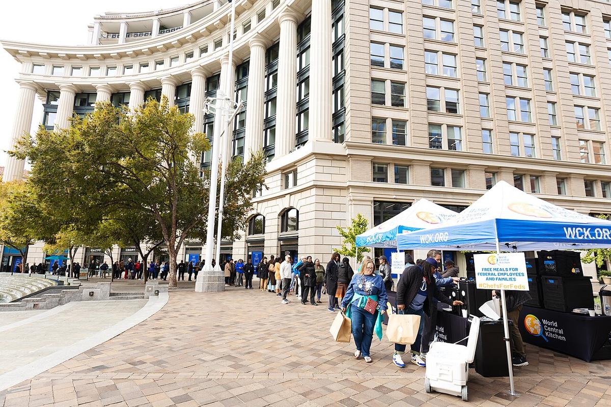 Long line for meals as World Central Kitchen serves meals to federal workers at the United States Navy Memorial plaza in Washington, D.C on October 31, 2025. The line stretches from two tents in the right foreground all the way to the leftmost edge of the picture, following the curve of a large building with huge columns in front.