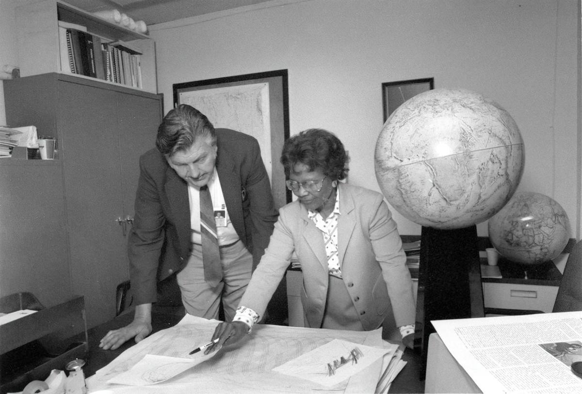 Gladys West and Sam Smith look over data from the Global Positioning System, which Gladys helped develop at the Naval Proving Ground in Dahlgren, VA in 1985. The two are seen poring over papers and charts in a crowded office with several globes and maps in the background.