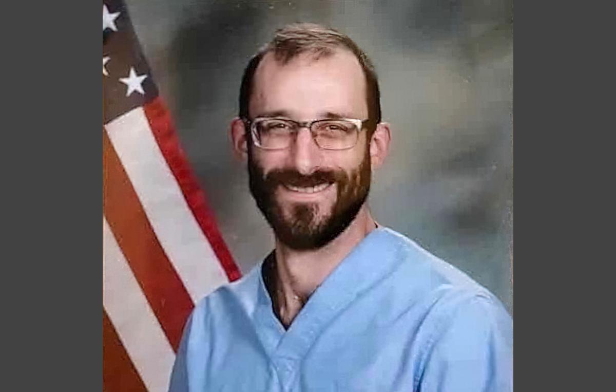 Alex Pretti VA Image (official portrait by United States Department of Veterans Affairs) The image shows a smiling, bearded young man wearing blue nurse's scrubs and glasses sitting in front of a flag.
