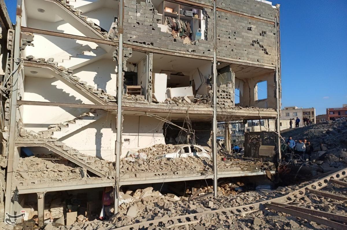 Bombing of a residential building in the city of Sanandaj, Iran. The image shows a multi-story center block building with one wall completely falling away, leaving the interior, Which is covered with rubble and debris, visible.