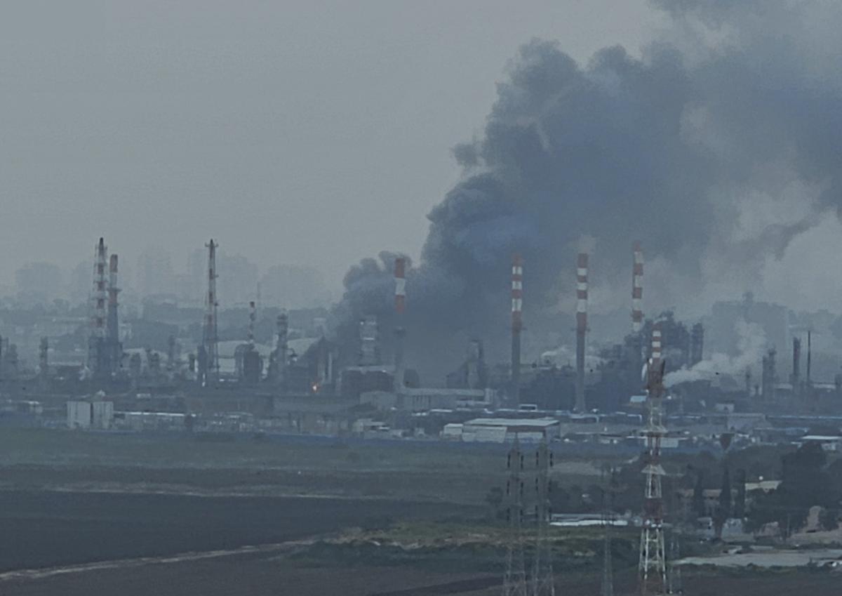 Missile interception fragments of an Iranian missile hit Haifa oil refineries in Haifa Bay, March, 19 2026 The strike caused temporary power outages after hitting a high-voltage line. The image shows an industrial area with multiple smoke stacks. Thick black smoke is rising from an impact site on the ground. The air everywhere in sight is a dirty gray. City buildings are just visible through the haze in the background.