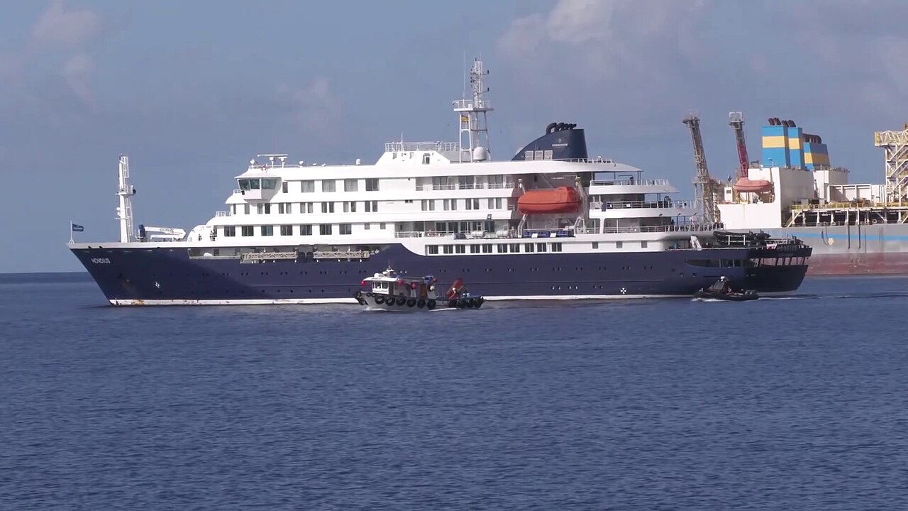 A large cruise ship, the MV "Hondius," anchored in a port with industrial structures visible in the background under a partly cloudy sky. A smaller vessel is visible in the foreground.