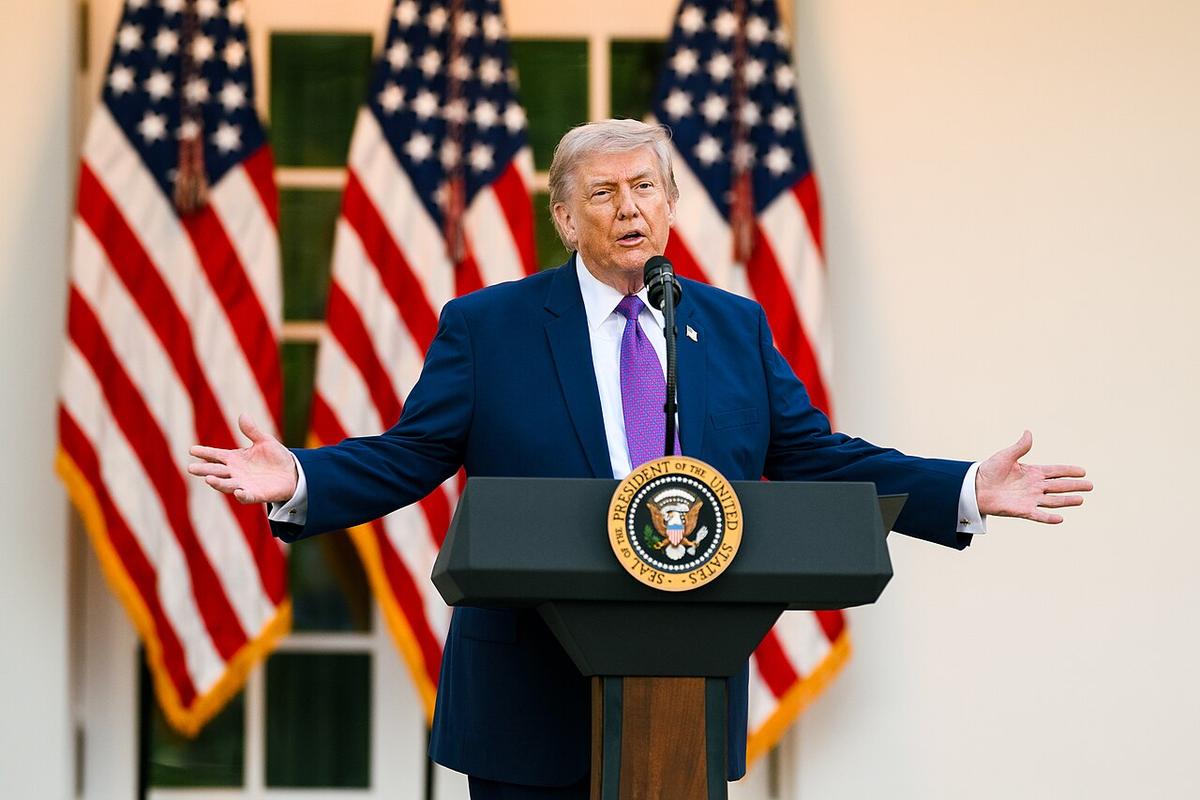 President Donald Trump is seen with his arm spread wide, speaking at a podium in front of three American flags.