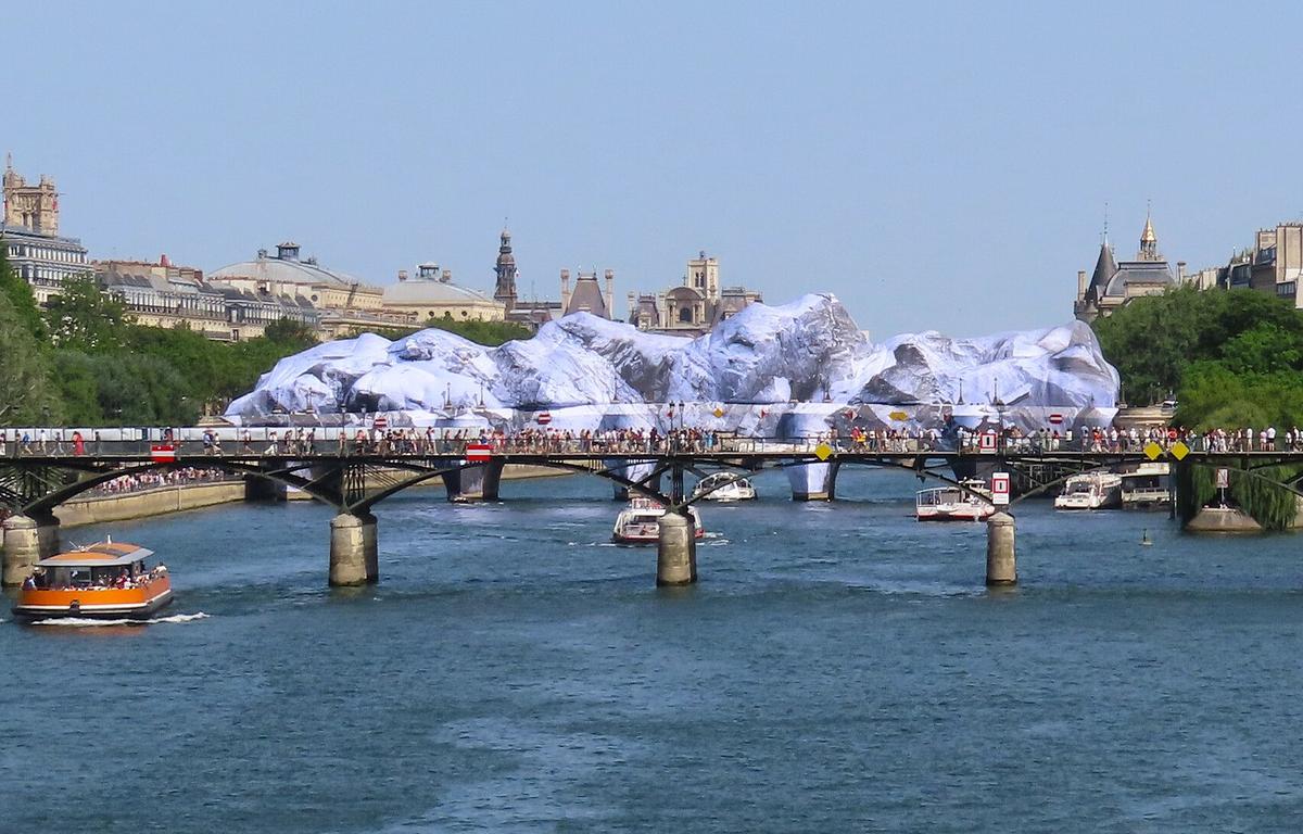 The Cave of Pont Neuf in Paris, a temporary art installation by JR. An inflated structure resembling mountains is seen in the middle of the Pont Neuf bridge as tourist boats approach it from either side.