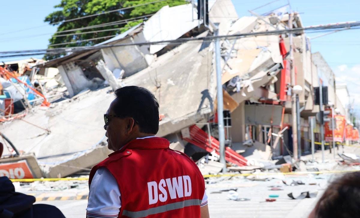 A collapsed concrete building with twisted metal and debris in the background, with a person wearing a red "DSWD" vest standing in the foreground at the scene of the damage.