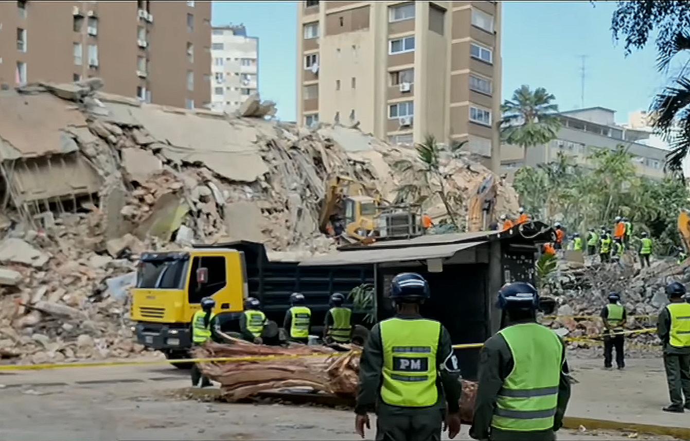 Emergency responders and heavy machinery work amidst the rubble of collapsed, heavily damaged concrete buildings in an urban area.
